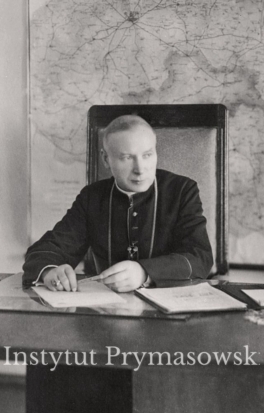 Bp.S.Wyszynski siedzi przy biurku nad papierami.na scianie wisi duza mapa .